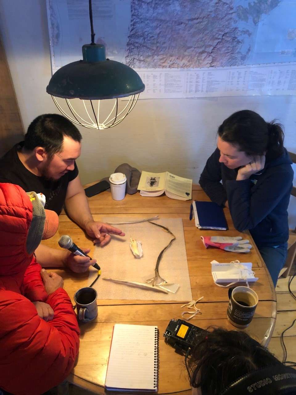 Trois personnes sont réunis autour d'une table, vue de haut. Un objet est posé au milieu. A gauche, deux inuit, dont l'un tient un micro, parlent de cet objet et le pointent. A droite, une personne observe. Une enregistreuse est sur la table.
