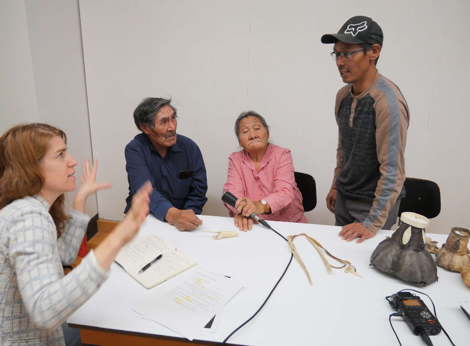 Quatre personnes sont réunis autour d'une table, sur laquelle sont posés des objets inuit et une enregistreuse. Trois personnes sont assises et une est debout. Une ainée inuk tient le micro. Deux autres personnes discutent.