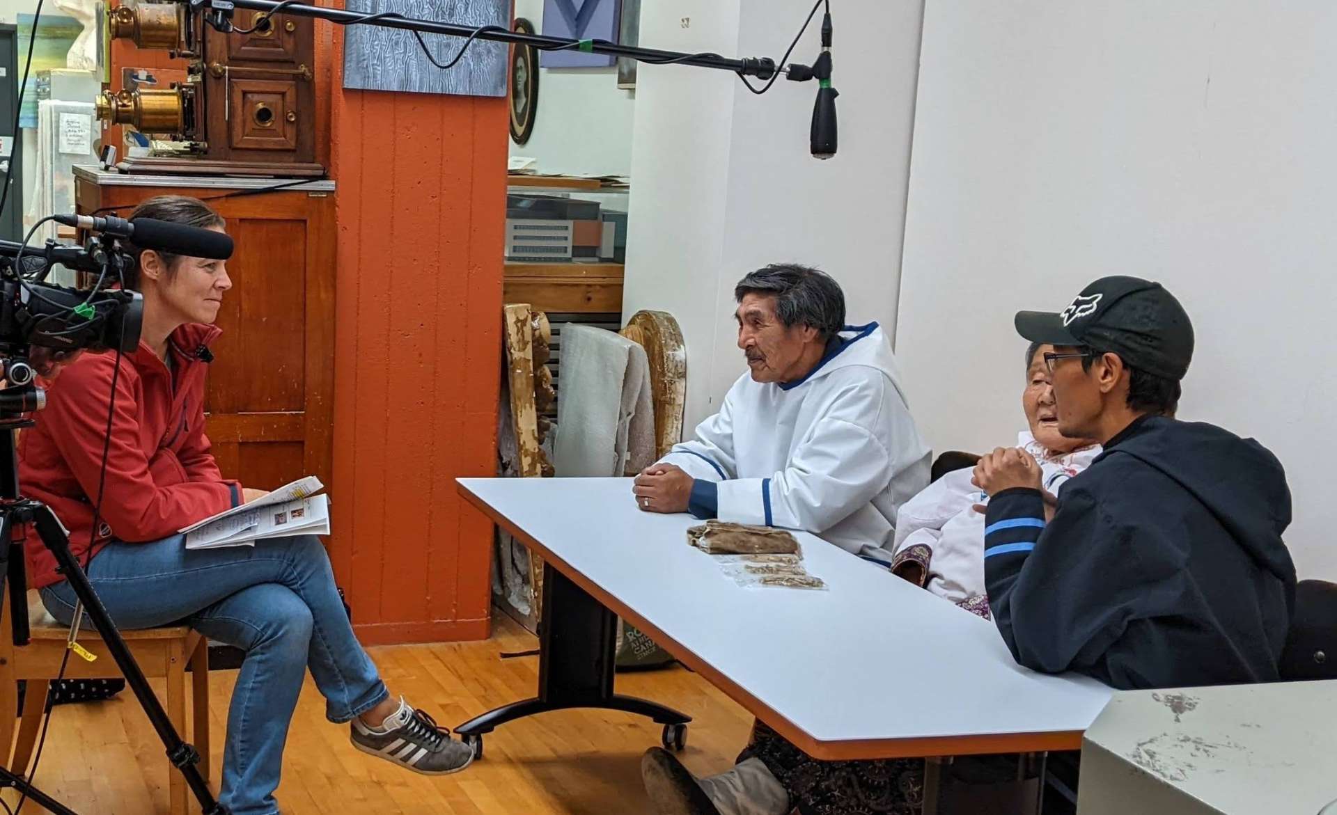 A droite de la photo, de profil, trois personnes sont assises devant une table sur laquelle est posé un objet. Ils partagent leur savoir sur celui-ci. Face a eux, une autre personne (assise), une caméra et un micro enregistrent les échanges.