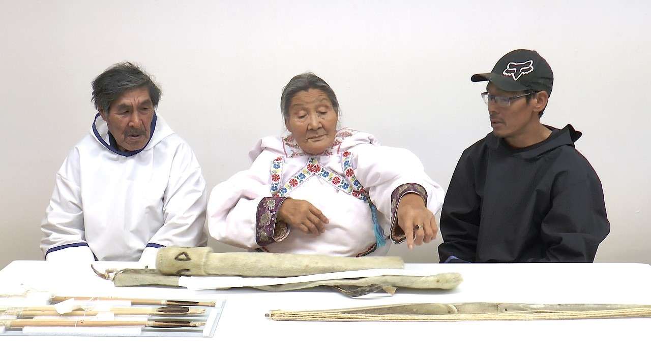 Deux ainés et un interprète inuit partagent leur savoir sur des objets posés sur une table devant eux. Ils sont tous assis et font face à l'appareil photo. Chaque ainé porte un amauti blanc avec de fines décorations de couleurs. Sur la table devant eux se trouvent cinq flèches, deux carquois et un arc que l'un des ainés pointe du doigt.