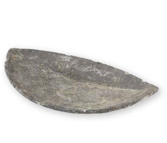 Lampe à l'huile en roche noircie de forme demi-circulaire, mais irrégulière, avec un creux central. La surface de la lampe montre des traces d'usure et des aspérités.