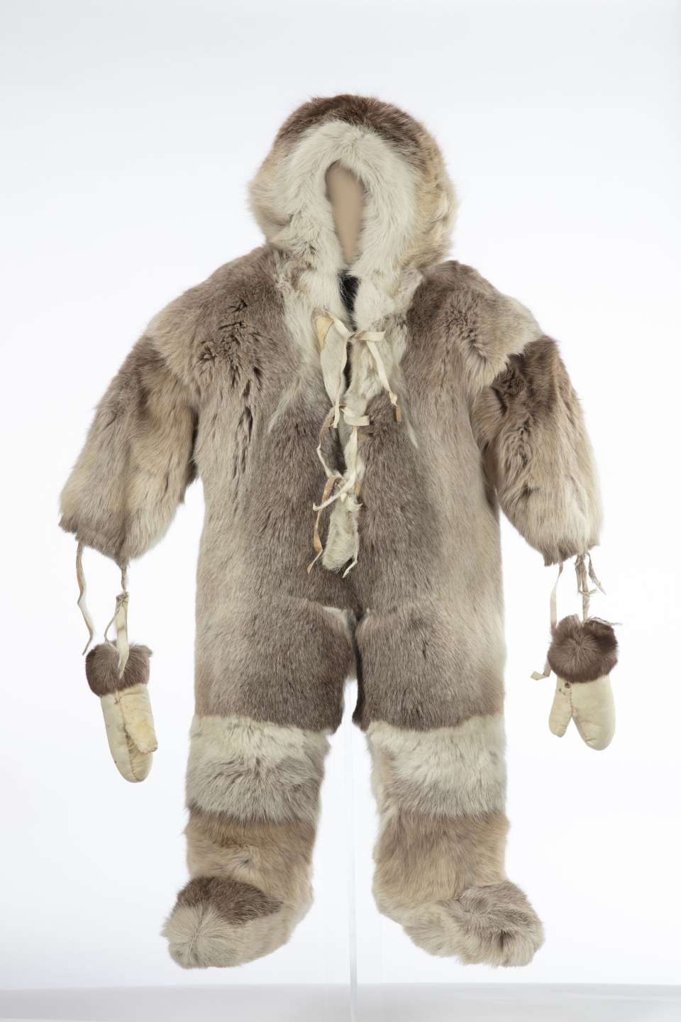 Combinaison en fourrure, vue de face, avec un capuchon. Des mitaines de cuir retourn&eacute;, orn&eacute;es de fourrure brune aux poignets, sont suspendues aux manches par des lani&egrave;res. La fourrure est de couleur plus claire autour du capuchon et au-dessus des bottes qui sont int&eacute;gr&eacute;es &agrave; la combinaison. La combinaison est ferm&eacute;e par un lacet &agrave; l&rsquo;avant au niveau de la poitrine et du cou.