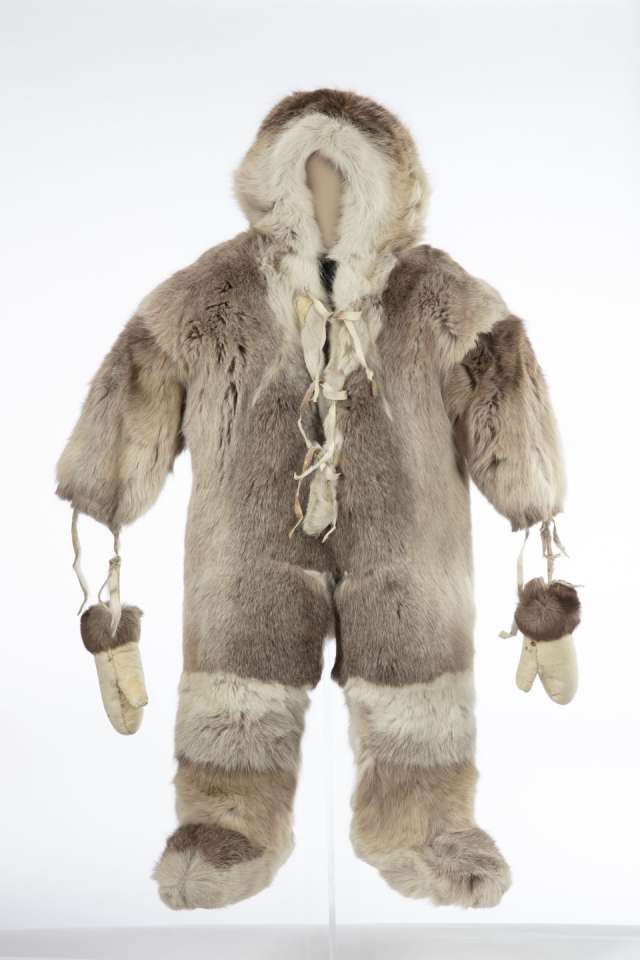 Combinaison en fourrure, vue de face, avec un capuchon. Des mitaines de cuir retourn&eacute;, orn&eacute;es de fourrure brune aux poignets, sont suspendues aux manches par des lani&egrave;res. La fourrure est de couleur plus claire autour du capuchon et au-dessus des bottes qui sont int&eacute;gr&eacute;es &agrave; la combinaison. La combinaison est ferm&eacute;e par un lacet &agrave; l&rsquo;avant au niveau de la poitrine et du cou.