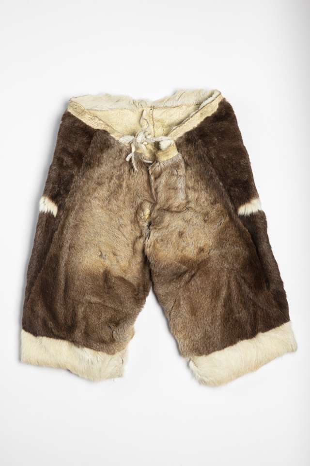 Pantalon en fourrure brune, vu de face, avec deux courtes bandes blanches sur les c&ocirc;t&eacute;s et une bande de fourrure blanche au bas de chaque jambe. La taille en cuir retourn&eacute; est ferm&eacute;e par un cordon nou&eacute; &agrave; l&rsquo;arri&egrave;re, mais visible &agrave; l&rsquo;avant.