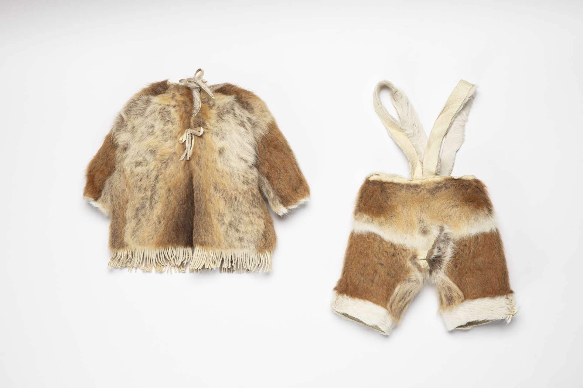 Petit ensemble en fourrure, vu de derri&egrave;re, compos&eacute; d&rsquo;un manteau et d&rsquo;un pantalon &agrave; bretelles. Le manteau est dot&eacute; de franges en bas et de cordons nou&eacute;s au niveau de l&rsquo;encolure. Une bande lat&eacute;rale de fourrure blanche est visible au milieu du pantalon, ainsi qu&rsquo;au niveau de la taille et du bas de chaque jambe. Le pantalon est dot&eacute; de larges bretelles en peau.