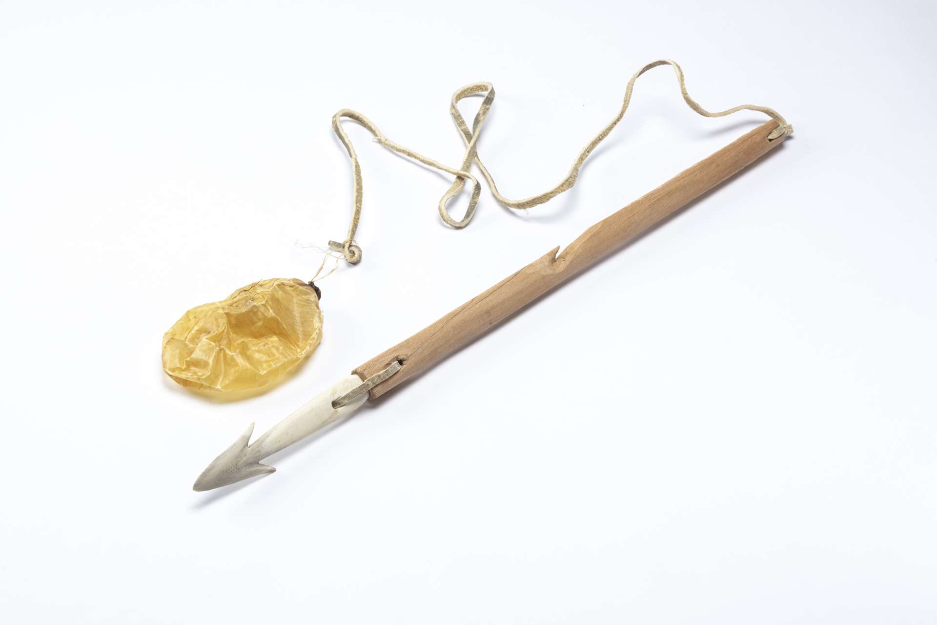 Harpon en bois avec une pointe en mati&egrave;re osseuse blanche, reli&eacute;e par une lani&egrave;re de cuir. Une autre lani&egrave;re de cuir est attach&eacute;e &agrave; un petit flotteur en forme de poche gonflable, jaun&acirc;tre.