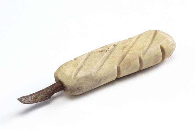 Outil avec un manche en mati&egrave;re osseuse blanche, grav&eacute; de lignes diagonales profondes et r&eacute;guli&egrave;res. Une lame m&eacute;tallique courb&eacute;e et us&eacute;e est fix&eacute;e &agrave; une extr&eacute;mit&eacute; du manche, montrant des signes de rouille.