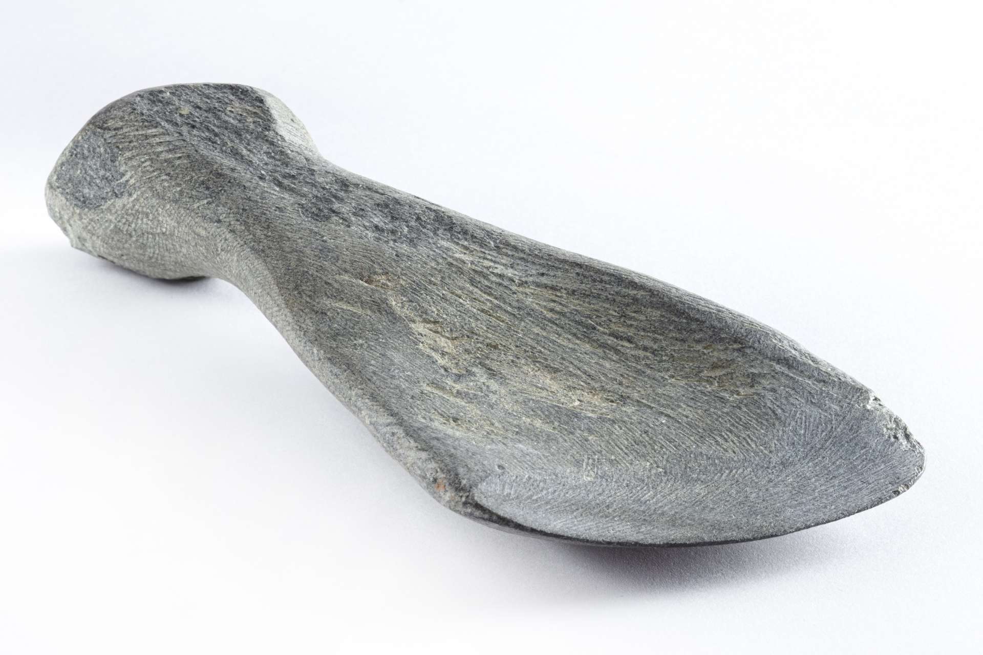Grattoir en roche grise sculpt&eacute;e, de forme allong&eacute;e et incurv&eacute;e. La surface pr&eacute;sente des lignes de sculpture et une l&eacute;g&egrave;re usure visible sur les bords arrondis.
