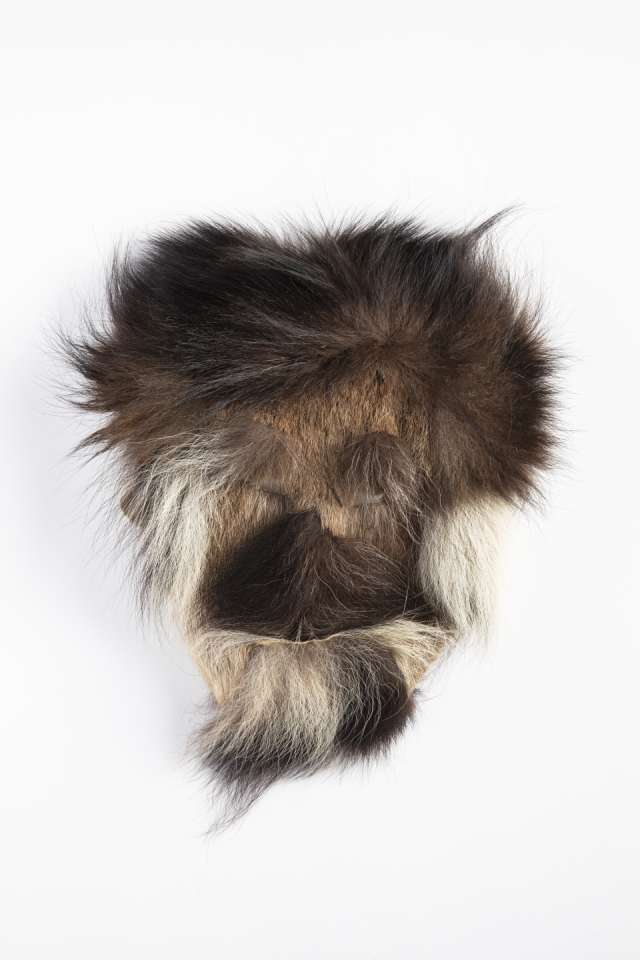 Masque en fourrure brune, vu de face, orn&eacute; de fourrure sombre, avec des touffes de fourrure noire et brune de part et d&rsquo;autre, et des zones plus claires sous le menton et autour des joues. La fourrure forme des lignes distinctes cr&eacute;ant une impression de cheveux, de barbe et de moustaches.