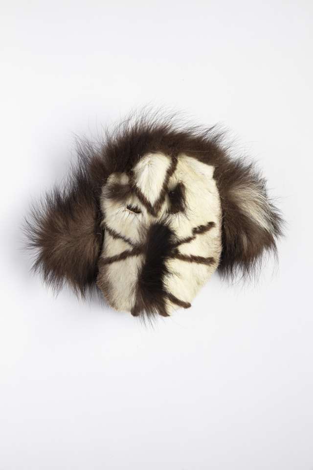 Masque en fourrure blanche, vu de face, avec un contraste marqu&eacute; entre les sections brunes, simulant des tatouages faciaux. Le nez, les yeux et les sourcils sont accentu&eacute;s par des touffes de fourrure plus sombre.
