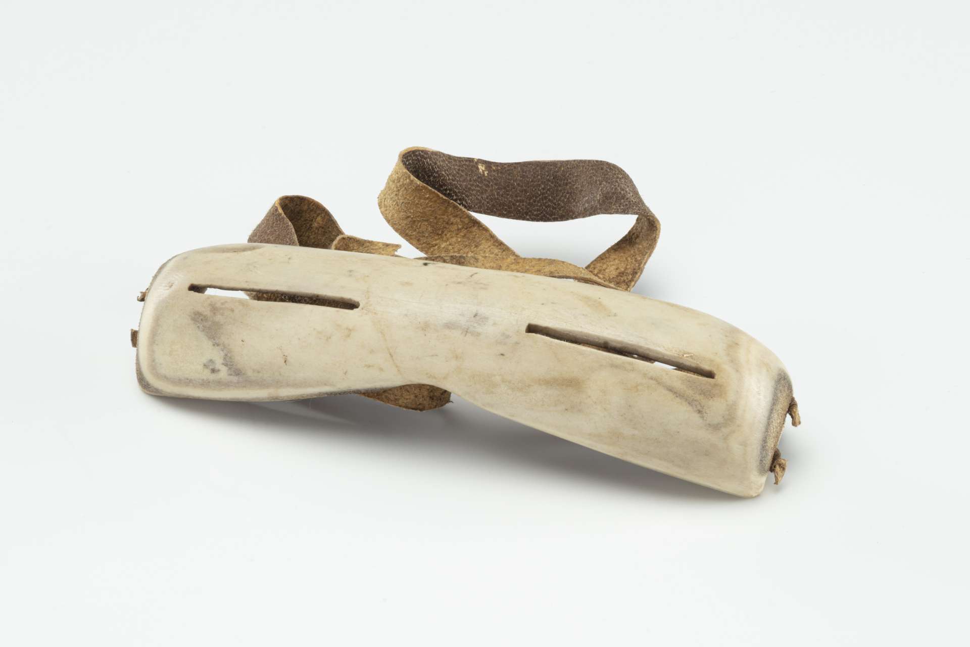 Lunettes sculpt&eacute;es dans une mati&egrave;re osseuse blanche, vues de devant, avec deux fentes &eacute;troites pour les yeux et une sangle en cuir brun.