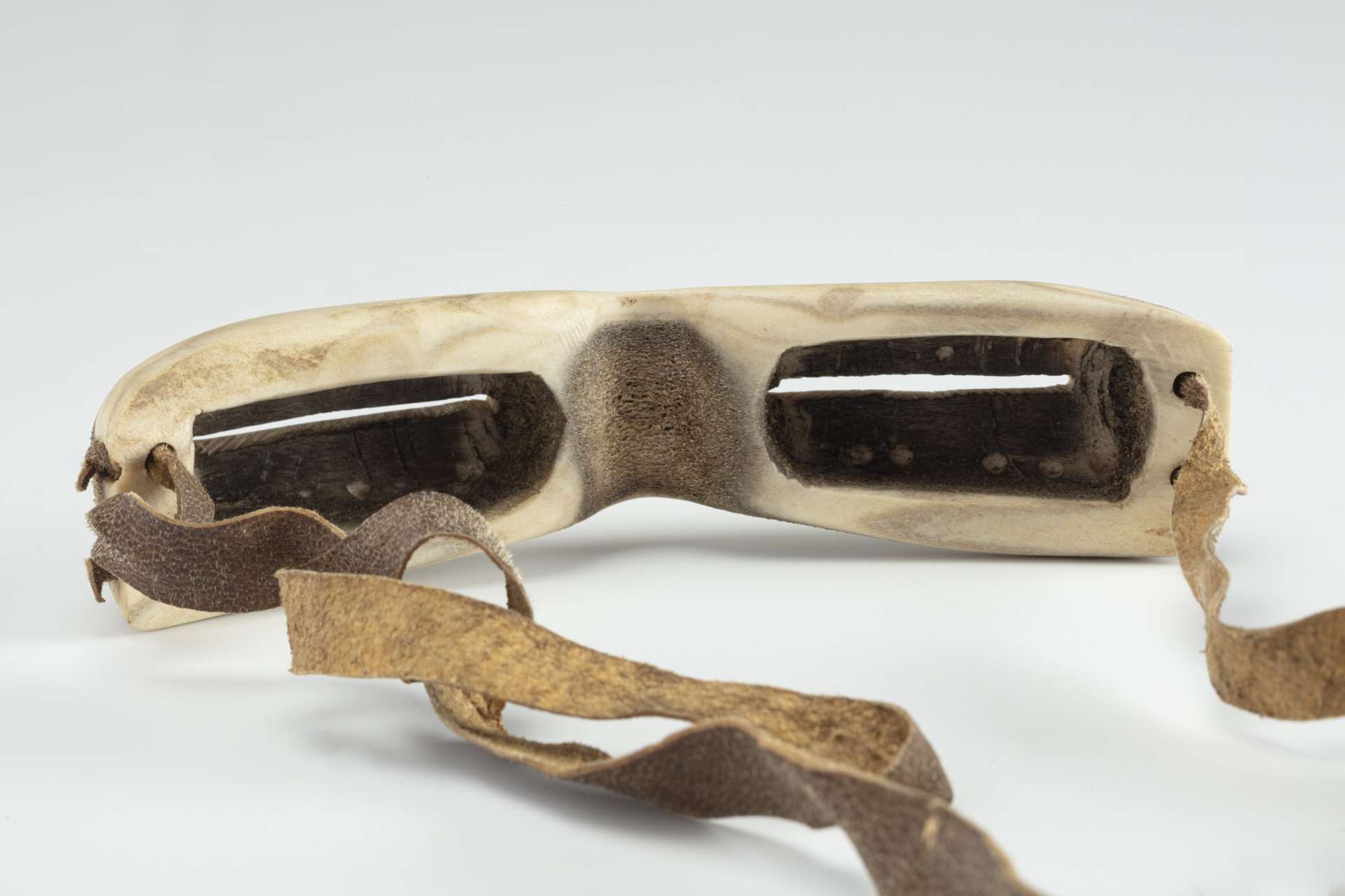 Lunettes sculpt&eacute;es dans une mati&egrave;re osseuse blanche, vues de derri&egrave;re, montrant l&rsquo;int&eacute;rieur creus&eacute; pour le nez et les yeux, avec une teinte fonc&eacute;e &agrave; l&rsquo;int&eacute;rieur et une sangle en cuir brun, fix&eacute;e aux extr&eacute;mit&eacute;s.