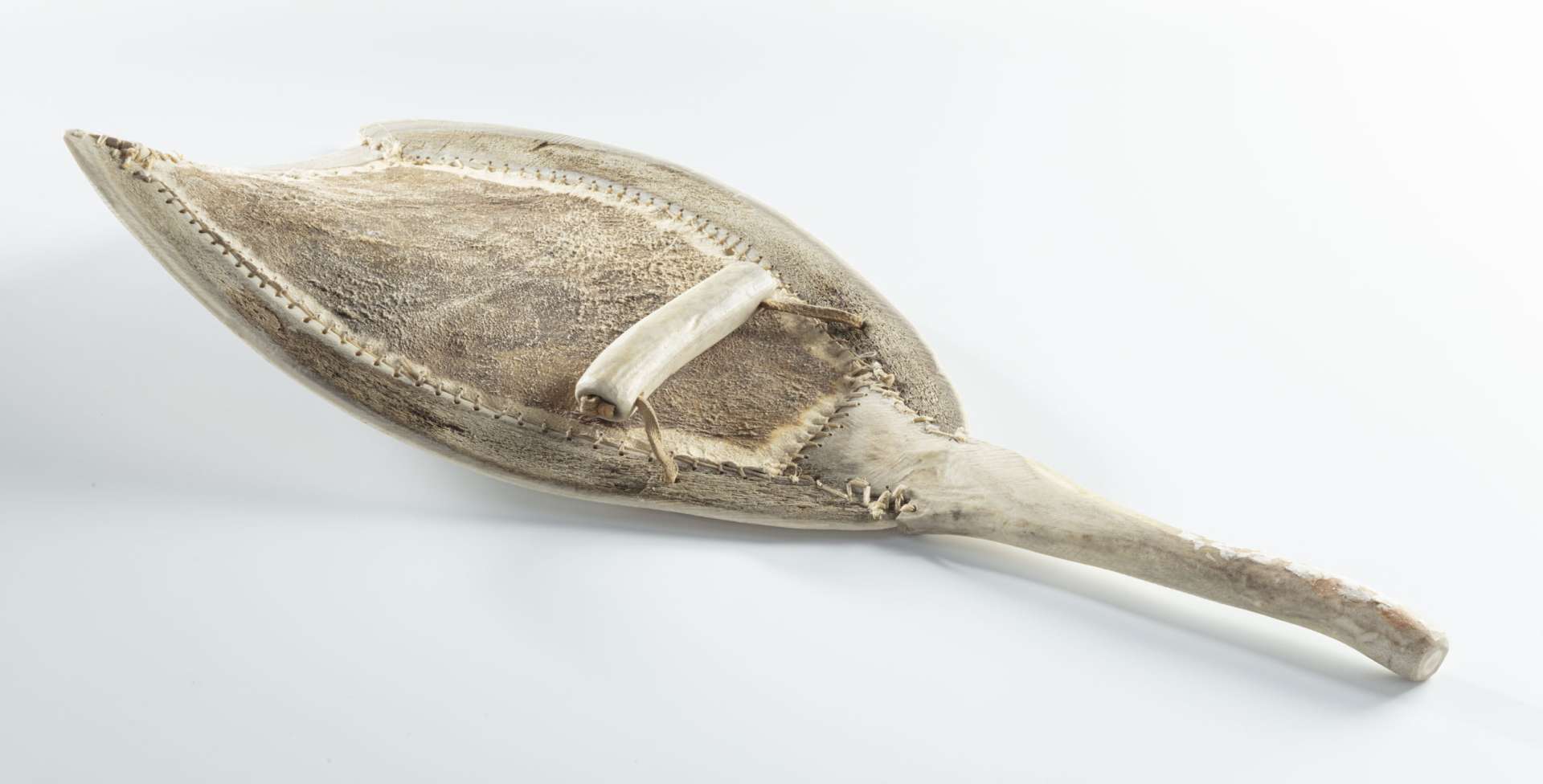 Pelle en mati&egrave;re osseuse blanche, vue de face, montrant le manche l&eacute;g&egrave;rement incurv&eacute;. Le fond en cuir, beige d&rsquo;un c&ocirc;t&eacute; et brun fonc&eacute; de l&rsquo;autre, est cousu le long des bords de l&rsquo;armature. Une poign&eacute;e en mati&egrave;re osseuse blanche, attach&eacute;e par une courroie de cuir, est positionn&eacute;e au centre de la pelle.