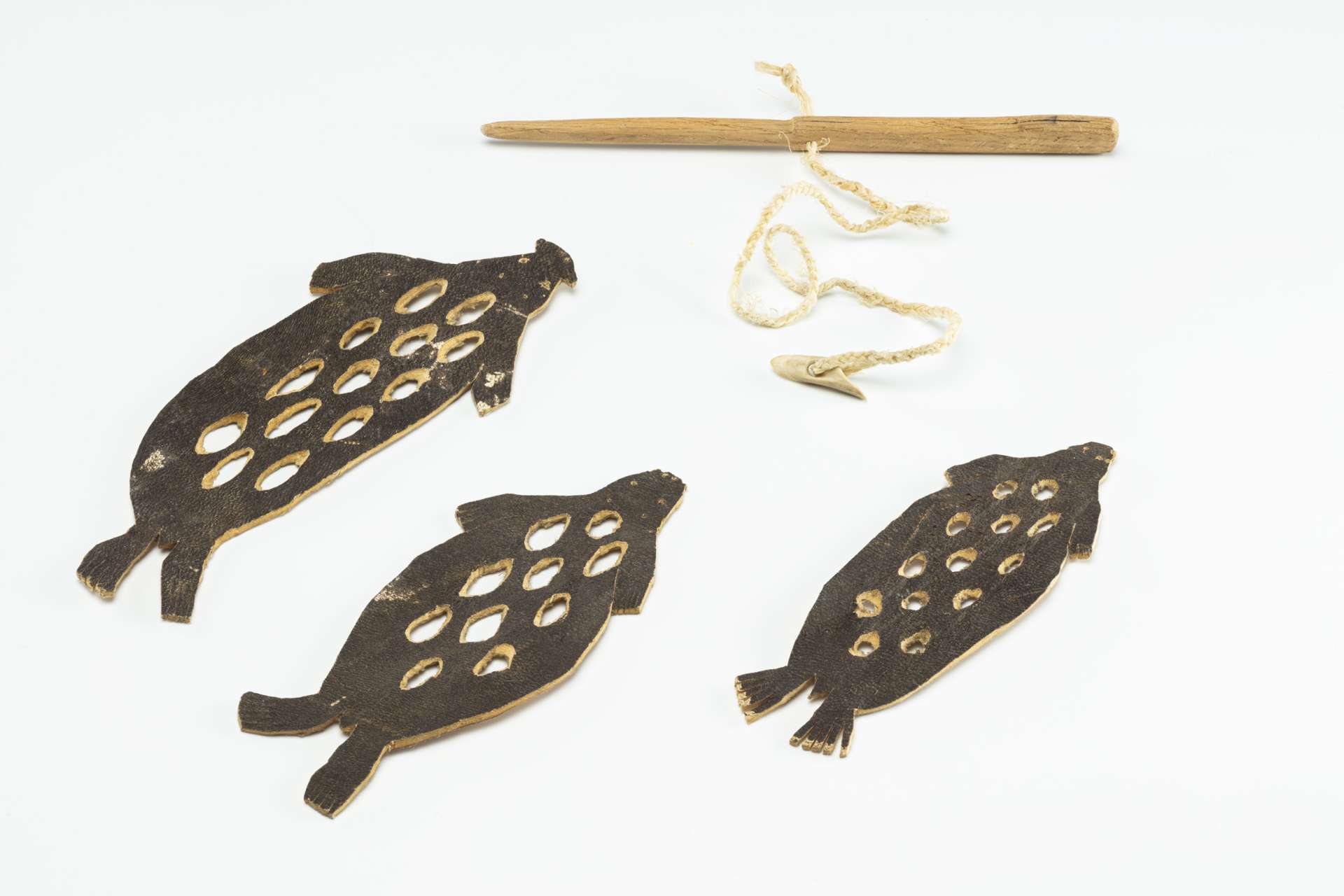 Trois morceaux de cuir brun d&eacute;coup&eacute;s en forme de phoque sont dispos&eacute;s c&ocirc;te &agrave; c&ocirc;te, chacun pr&eacute;sentant des perforations circulaires. &Agrave; c&ocirc;t&eacute; figurent un petit harpon en bois avec une ligne tress&eacute;e et une t&ecirc;te de harpon en os.