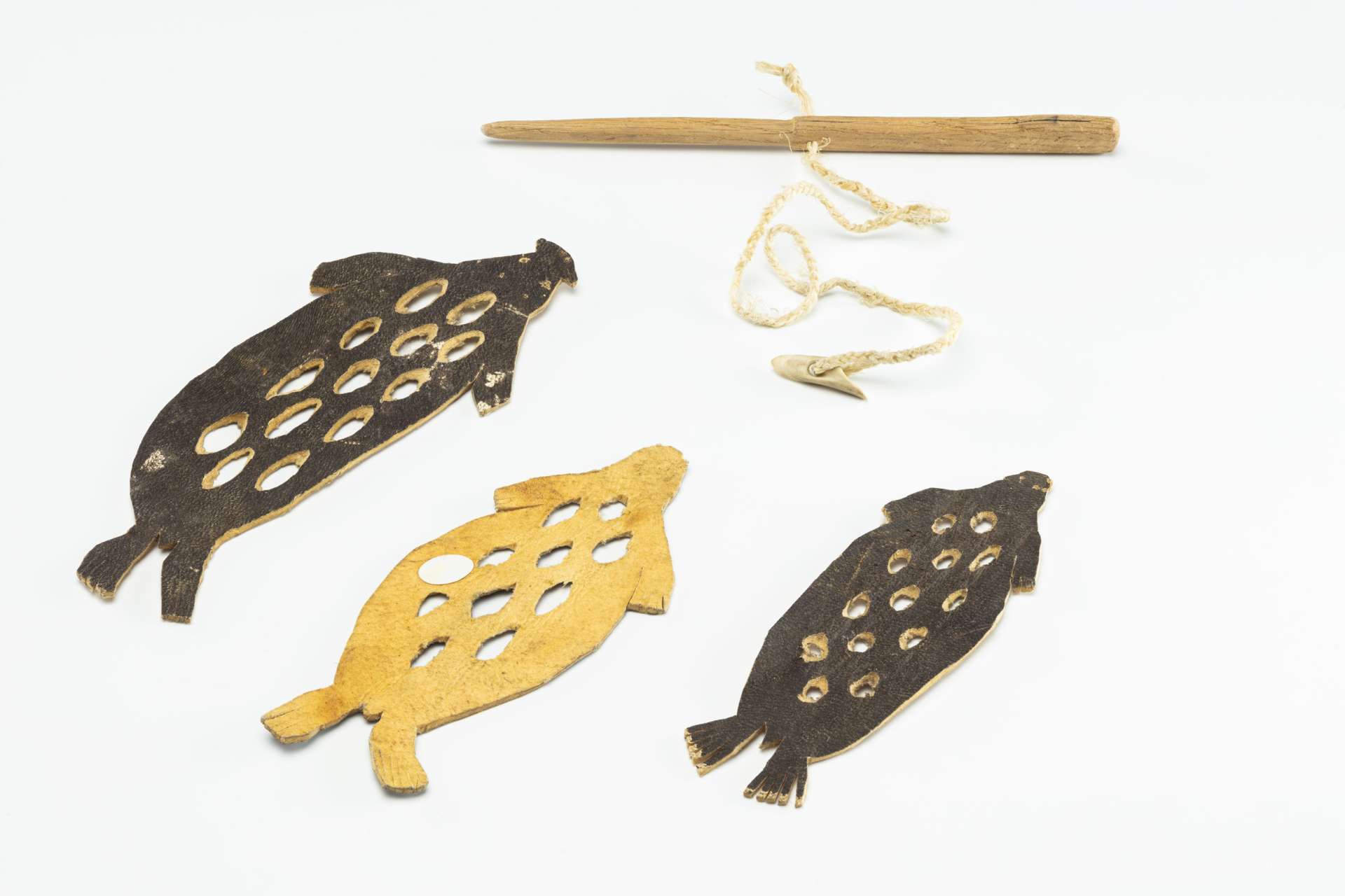 Trois morceaux de cuir d&eacute;coup&eacute;s en forme de phoque, perfor&eacute;s de trous circulaires, sont dispos&eacute;s c&ocirc;te &agrave; c&ocirc;te. Deux sont pr&eacute;sent&eacute;s c&ocirc;t&eacute; cuir brun fonc&eacute; et le troisi&egrave;me, au milieu, c&ocirc;t&eacute; su&egrave;de brun clair. &Agrave; c&ocirc;t&eacute; figurent un petit harpon en bois avec une ligne tress&eacute;e et une t&ecirc;te de harpon triangulaire en os.