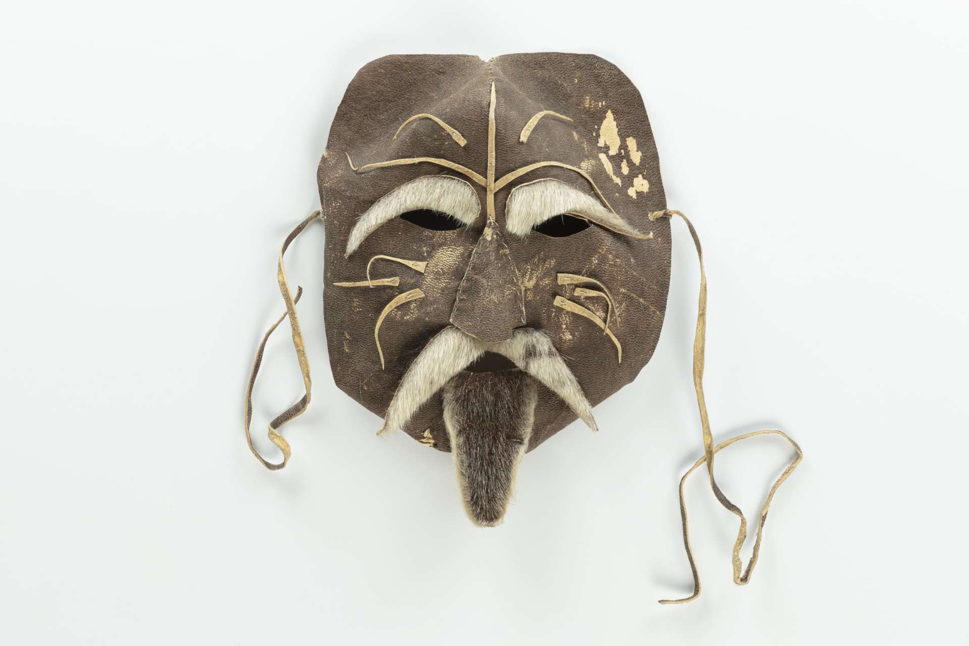 Masque en cuir brun, vu de face, avec des ouvertures pour les yeux et des sourcils, une moustache et une barbichette en fourrure. Le masque est orn&eacute; de motifs de cuir en relief et est muni de cordons en cuir pour l&rsquo;attacher.