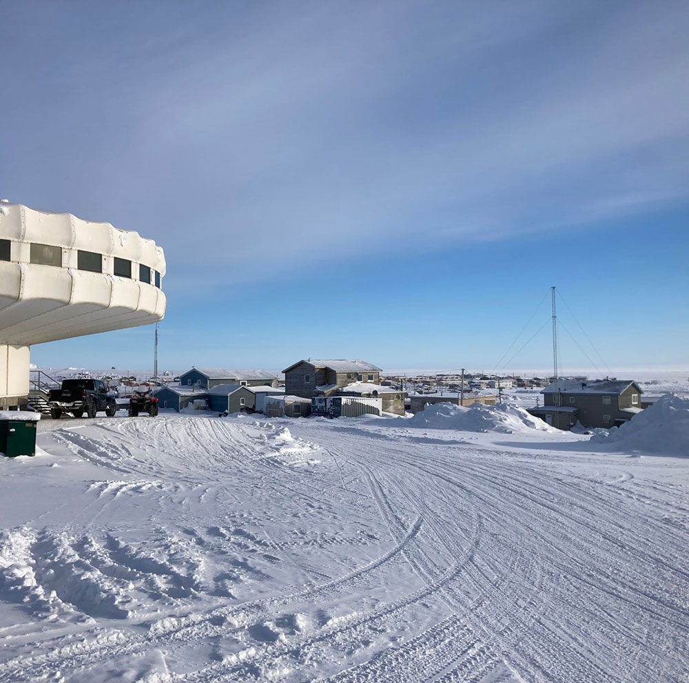 Cette photo illustre Igloolik en hiver : il y a de la neige au sol, un batiment en forme de champignon sur la gauche, dont seul la moitié apparait, et plusieurs maisons en arrière plan.